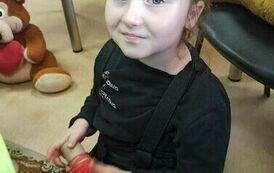 Dziewczynka na czarno ubrana siedzi przed zielonym krzesełkiem trzymając w ręku motek czerwonych nici.