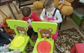 Chłopiec z dziewczynką trzymają w ręku motki z nićmi. Przed nimi stoją małe zielone krzesełka na kt&oacute;rych na oparciach są przyczepione twarze misi&oacute;w z dziurką zrobioną w ustach przez kt&oacute;re przewleczone są nici. Na siedzeniach są talerzyki kolorowe z nitkam