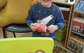 Chłopiec trzyma w ręku motek z nićmi. Przed nim stoi małe zielone krzesełko na kt&oacute;rym na oparciu jest przyczepiona twarz misia z dziurką zrobioną w ustach przez kt&oacute;rą przewleczona jest nić. Na siedzeniu jest czerwony talerzyk.