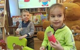 Chłopiec i dziewczynka trzymają w ręku motki z nićmi. Przed nimi stoją małe zielone krzesełka na kt&oacute;rych na oparciach są przyczepione twarze misi&oacute;w z dziurką zrobioną w ustach przez kt&oacute;re przewleczone są nici. 