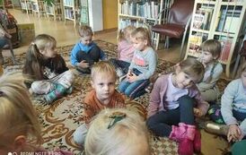 Dzieci siedzą na dywanie i słuchają bajki czytanej przez bibliotekarkę.