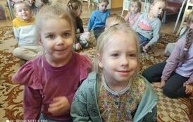 Dzieci siedzą na dywanie, z przodu dobrze widoczne dwie dziewczynki o blond włosach.