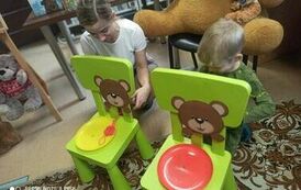 Dziewczynka i chłopiec trzymają w rękach motki nici. Stoją przed małymi zielonymi krzesełkami na kt&oacute;rych oparciach sa naklejone twarze misia z dziurką w ustach przez kt&oacute;rą przełożona jest nic. Na siedzeniach są kolorowe miski, na jednej jest rozwinięta ni