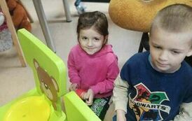 Chłopiec i dziewczynka trzymają w rękach motki nici. Stoją przed małymi zielonymi krzesełkami na kt&oacute;rych oparciach sa naklejone twarze misia z dziurką w ustach przez kt&oacute;rą przełożona jest nic. Na siedzeniach są kolorowe miski.