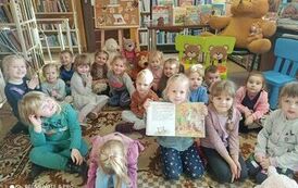 Dzieci siedzą na dywanie, jedna z dziewczynek ma otwarte książkę na bajce o misiach i ją pokazuje.