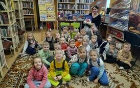 Zdjęcie grupowe dzieci siedzących na dywanie, z tyłu za nimi siedzi bibliotekarka trzymając książkę w rękach.
