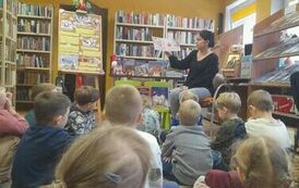 Dzieci siedzą na dywanie odwr&oacute;cone tyłem i patrzą w stronę bibliotekarki, kt&oacute;ra czyta im bajkę.