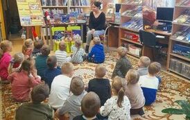 Dzieci siedzą na dywanie odwr&oacute;cone tyłem i patrzą w stronę bibliotekarki, kt&oacute;ra  im o czymś opowiada.