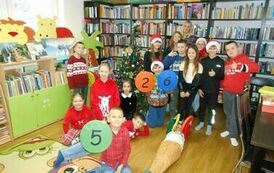 Grupa dzieci stojących ze swoją Panią przy przystrojonej choince. Niekt&oacute;re dzieci trzymają w ręku siedziska z numerkami.