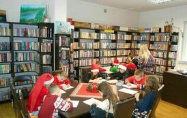 Dzieci siedzą przy stołach i robią świąteczne karty pracy. Obok stoi ich Pani. Niekt&oacute;re dzieci mają ubrane czapki mikołajkowe.