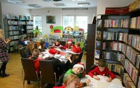 Dzieci siedzą przy stołach i robią świąteczne karty pracy. Obok stoi ich Pani. Niekt&oacute;re z dzieci mają ubrane czapki mikołajkowe.