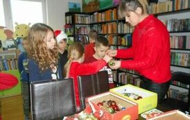 Na stole leżą ozdoby choinkowe, stoi przy nich bibliotekarka i podaje dzieciom ozdoby do strojenia choinki.