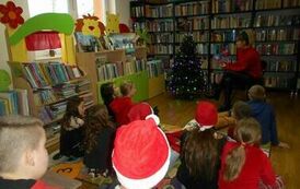 Dzieci siedzą na dywanie i patrzą w stronę bibliotekarki, kt&oacute;ra czyta im bajkę. Z boku stoi oświetlona choinka.