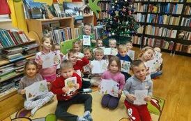 Dzieci siedzą na dywanie przy choince i trzymają w rękach swoje zrobione kartki świąteczne.