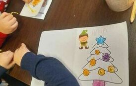 Na stole widać rączki dzieci robiące kartki świąteczne za pomocą kredek, naklejek, brokatu i cekink&oacute;w.Widać przystrojoną na kartce choinkę.