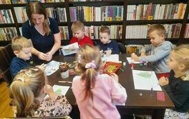 Dzieci siedzą przy stole i robią kartki świąteczne przy pomocy kredek, naklejek, brokatu i cekink&oacute;w. Nad nimi pochylona jest Pani i patrzy jak to wykonują.