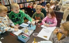 Dzieci siedzą przy stołach i robią kartki świąteczne przy pomocy kredek, naklejek, brokatu i cekink&oacute;w.