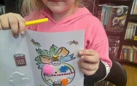 Dziewczynka pokazuje swoją zrobioną kartkę świąteczną. Widać na niej przystrojoną bombkę.