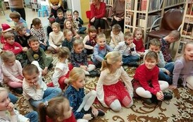 Dzieci siedzą na dywanie, a z tyłu na krzesełkach siedzą opiekunowie.