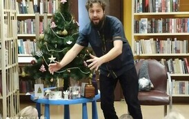 Szymon G&oacute;ralczyk pokazuje rękoma coś dzieciom. Obok stoi na stoliczku przystrojona choinka.