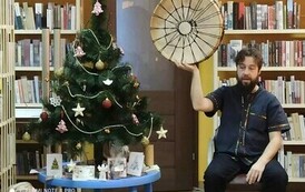 Po lewej stronie stoi na stoliczku choinka i obok niej życzenia. Po prawej stronie siedzi Szymon G&oacute;ralczyk i trzyma uniesione do g&oacute;ry drewniane koło owinięte sznurkami.