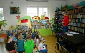 Dzieci siedzą na dywanie i patrzą w stronę bibliotekarki, kt&oacute;ra im coś opowiada. W tle widać choinkę.