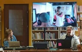 Z przodu przy stolikach siedzą artyści z rozłożynym sprzętem i patrzą na ekran na kt&oacute;rym jest wyświetlany filmik z nagrywania muzyki przez młodzież.