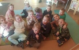 Grupa dzieci siedzi na dywanie i słucha bajki, niekt&oacute;re dzieci usmiechają się do zdjęcia