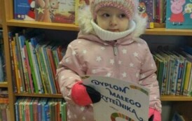 Mała dziewczynka trzyma w rączce sw&oacute;j dyplom. Za nią widać p&oacute;łki z książkami.