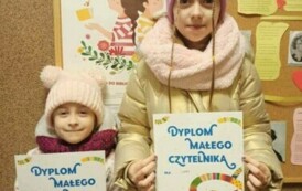Dwie dziewczynki pokazują swoje dyplomy. Za nimi jest metalowa tablica z plakatem promującym akcję Mała Książka Wielki Człowiek i z innymi ciekawostkami kulturowymi.
