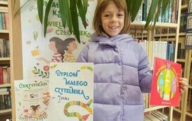 Uśmiechnięta dziewczynka w fioletowej kurtce trzyma w rękach dyplom i wypożyczoną książeczkę. Obok stoi tablica korkowa z plakatem promującym akcję Mała Książka Wielki Człowiek.