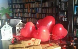 Na stoliku są wyłożone książki zapakowane w papier przystrojony serduszkami, obok są balony, lampiony i serduszka.