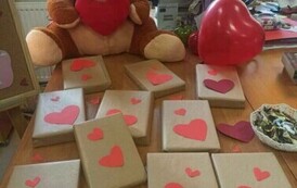 Na biurku siedzi miś z sercem na brzuchu i leżą książki opakowane w papier i przystrojone serduszkami i są balony czerwone.