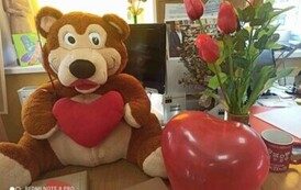 Na biurku siedzi miś z sercem na brzuchu i leżą książki opakowane w papier i przystrojone serduszkami i są balony czerwone i kwiatki w wazonie.