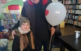 Dziewczynka stoi razem z czytelnikiem, kt&oacute;ry trzyma w ręce biały balon z serduszkiem.