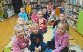 Dzieci siedzą na dywanie i słuchają bajki czytanej przez bibliotekarkę.