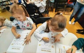 Z przodu dwie dziewczynki wykonują kartę pracy, w tle widać inne dzieci przy stołach.