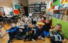 Cała grupa dzieci siedzi na dywanie i słucha bajki czytanej przez bibliotekarkę. Za nimi na krzesełku siedzi nauczycielka.