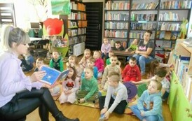 Dzieci siedzą na dywanie i słuchają bajki, kt&oacute;rą czyta im bibliotekarka.