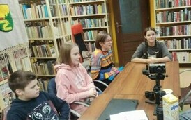 Młodzież siedzi przy stole w bibliotece, na stole stoi kamera na stojaku i akcesoria do tego.