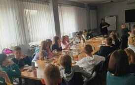 Przy stołach są rodzice i dzieci. Patrzą do przodu na Iwonę Skwarek, kt&oacute;ra im opowiada o warsztatach.