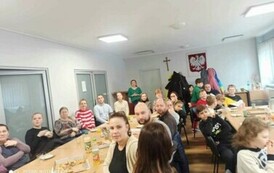 Przy stołach na białej sali siedzą licznie rodzice i ich dzieci oglądając nagrania filmowe i muzyczne dzieci. Na stołach jest pizza i napoje.