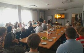 Dzieci i rodzice przy stołach na białej sali oglądają zmontowane filmiki na dużym ekranie na kt&oacute;rym widać ich pociechy.