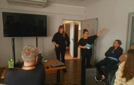 W białej sali z przodu widać kierowniczkę, kt&oacute;ra ma wyciągniętą rękę do g&oacute;ry, obok stoi Iwona, na przecwko dużego ekranu siedzi Piotr Martin i widać rodzic&oacute;w.