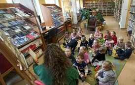 Dzieci siedzą na dywanie i patrzą w stronę bibliotekarki, kt&oacute;ra czyta im bajkę.