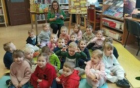 Dzieci siedzą w grupie na karimatach, z tyłu po środku siedzi bibliotekarka z książeczką o zajączku.