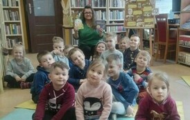 Dzieci siedzą w grupie na karimatach, z tyłu po środku siedzi bibliotekarka z dwiema książeczkami świątecznymi w ręku pokazując je.