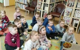 Dzieci siedzą na dywanie i patrzą w stronę bibliotekarki, kt&oacute;ra czyta im bajkę.