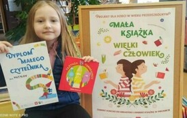 Dziewczynka w blond włosach trzyma w rekach dyplom i wypożyczoną książeczkę. z prawej strony stoi tablica z plakatem promującym akcję Mała Książka Wielki Człowiek.