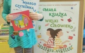 Uśmiechnięta dziewczynka trzyma w rękach dyplom, kartę czytelniczą i wypożyczoną książeczkę. Z prawej strony stoi korkowa tablica z plakatem promującym projekt czytelniczy.